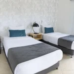 Una luminosa y moderna habitación de hotel en Cabañas Consotá cuenta con dos camas individuales con sábanas blancas, mantas grises y almohadas azules. Junto a cada cama hay pequeñas mesillas de noche de madera y lámparas negras, y una ventana llena el espacio de luz natural.