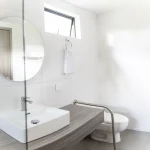 Moderno baño accesible en Cabañas Consotá con espejo redondo, lavabo cuadrado blanco sobre encimera de madera, toallero con toalla, barra de apoyo, inodoro y una pequeña ventana que deja entrar la luz natural.