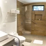 Moderno cuarto de baño accesible en Cabañas Consotá con ducha acristalada, azulejos de madera en la pared, barra de apoyo, toallas dobladas en un estante, inodoro blanco, lavabo, espejo redondo y una pequeña ventana que deja entrar la luz natural.