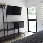 Dormitorio minimalista en Cabañas Consotá con un televisor montado en la pared, una estantería de metal negro con toallas dobladas, un cajón, una puerta de cristal que da al exterior, una ventana con persianas beige y una cama con una funda acolchada oscura.