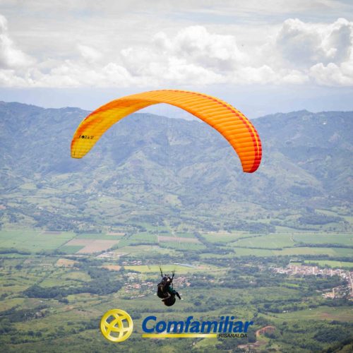 Una persona vuela en parapente con un paracaídas naranja y amarillo sobre un valle verde y montañas lejanas. Abajo, un logo dice Comfamiliar Risaralda con un símbolo circular amarillo, al lado de la Biblioteca Pública Municipal.