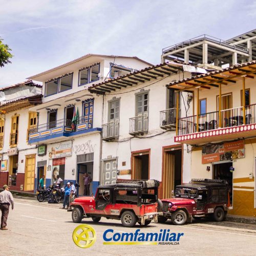 Coloridos edificios coloniales bordean una calle soleada con jeeps aparcados y algunas personas paseando. Un árbol da sombra a la izquierda, cerca de la Biblioteca Pública Municipal Isaías Naranjo, y un logotipo de Comfamiliar Risaralda es visible en la parte inferior central.