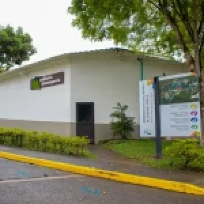 Un pequeño edificio blanco con un cartel en el que se lee Laboratorio Entomología se alza junto al Auditorio Barranquero, con un gran cartel informativo, un árbol y arbustos a lo largo de una acera húmeda en un exuberante entorno al aire libre.