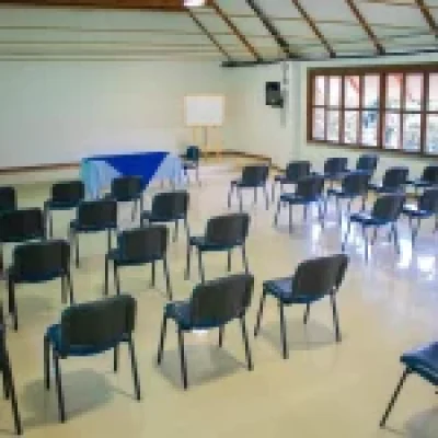 Una luminosa y vacía sala de conferencias del Auditorio Golondrina presenta hileras de sillas negras frente a una pequeña mesa y una pizarra blanca. Grandes ventanales llenan el espacio de luz natural bajo un techo alto e inclinado con vigas a la vista.