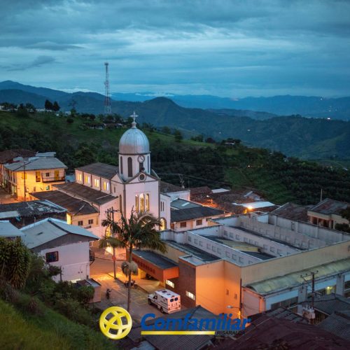 Una pequeña ciudad con una iglesia blanca en el centro, rodeada de colinas y montañas al atardecer. Luces cálidas iluminan las calles y los edificios. El logotipo de Comfamiliar Risaralda y la Biblioteca Pública son visibles en la parte inferior.