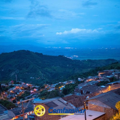 Vista panorámica nocturna de una ciudad montañosa con casas y luces dispersas, exuberantes colinas verdes al fondo, la Biblioteca Pública cerca y el logotipo de "Comfamiliar Risaralda" en la parte inferior de la imagen.