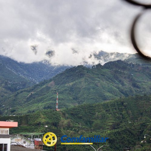 Exuberantes montañas verdes bajo un cielo nublado, con algunos edificios en primer plano, entre ellos la Biblioteca Pública Municipal Carlos Arturo Bolívar, y la palabra Comfamiliar con un logotipo amarillo en la parte inferior de la imagen.
