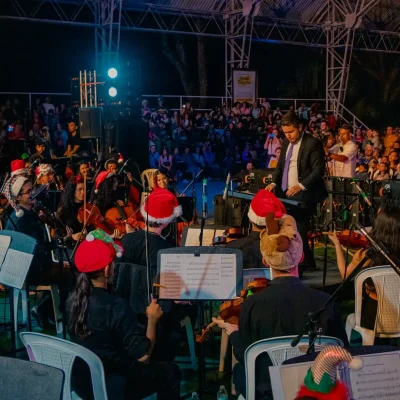 Una orquesta con sombreros de fiesta actúa en el escenario ante el público por la noche. El director se sitúa al frente, y el público se sienta apretujado en un Polideportivo bajo un toldo metálico, disfrutando de la música en este vibrante recinto al aire libre.