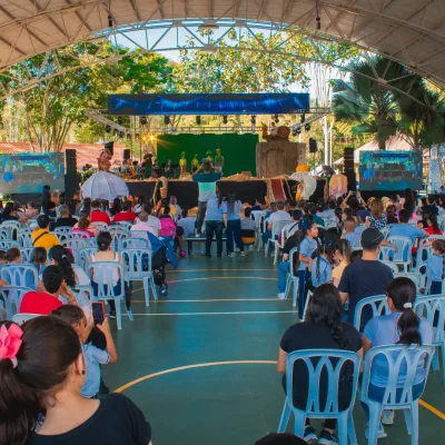 Una gran multitud se sienta en sillas de plástico frente a un escenario en el coliseo cubierto. Personas de todas las edades observan a los artistas bajo el recinto cubierto al aire libre, con árboles visibles al fondo.
