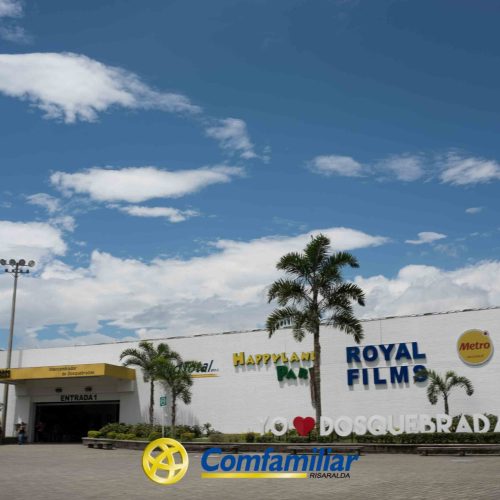 Un edificio blanco en Dosquebradas con carteles de Royal Films, Happyland y Comfamiliar, palmeras y un gran mural I ♥ Dosquebradas en el exterior bajo un cielo azul con nubes. La Biblioteca Pública cercana le añade encanto.