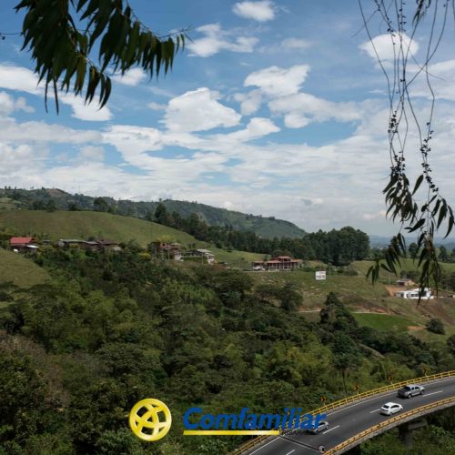 Una carretera en curva serpentea entre frondosas colinas verdes y casas dispersas bajo un cielo parcialmente nublado. En primer plano, el logotipo de Comfamiliar Dosquebradas aparece en la parte inferior, destacando su conexión con la cercana Biblioteca Pública.