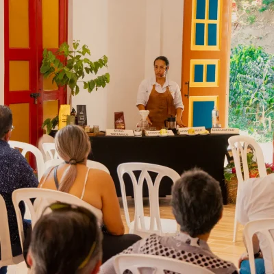 Una persona, de pie detrás de una mesa con varios objetos, habla a un público sentado en sillas blancas en una colorida sala de la Fonda Réplica de Pereira Antigua, con puertas y ventanas de vibrantes colores rojo y amarillo.