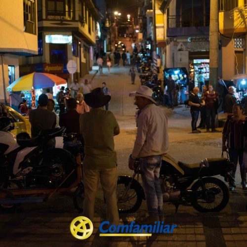 Animada escena callejera nocturna en Guática con gente socializando, motos aparcadas, vendedores ambulantes y tiendas iluminadas por luces artificiales. En primer plano, dos hombres con sombrero junto a motocicletas cerca de la Biblioteca Pública.