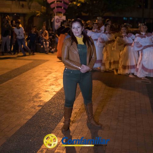 En primer plano, una mujer posa con confianza sobre un paseo nocturno de ladrillo, mientras un grupo de personas ataviadas con trajes tradicionales se reúnen detrás de ella. El logotipo Comfamiliar Guática aparece en la parte inferior, destacando la conexión con la Biblioteca Pública.