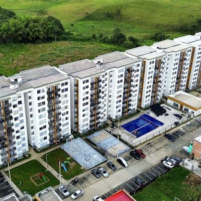 Vista aérea del Mirador Campestre, un moderno complejo de apartamentos con múltiples edificios blancos, una piscina central azul, canchas de baloncesto, zonas de aparcamiento y campos verdes circundantes.