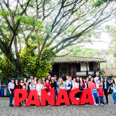 Un grupo de personas posa delante de grandes letras rojas "PANACA", con un edificio tradicional y frondosos árboles de fondo, bajo un cielo brillante y parcialmente nublado.