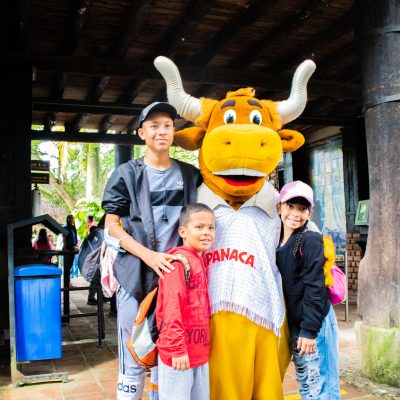 Tres niños sonríen junto a una persona disfrazada de vaca blanca y marrón con la etiqueta "PANACA", en un entorno al aire libre con paredes de ladrillo y vigas de madera.