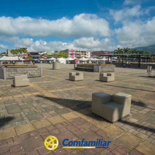 Una plaza amplia y abierta con bancos de hormigón y jardineras de piedra bajo un cielo azul salpicado de nubes. Edificios, carpas y palmeras en el fondo, cerca de la Biblioteca Pública Hugo Ángel Jaramillo. En la parte inferior aparece el logotipo de "Comfamiliar Risaralda".