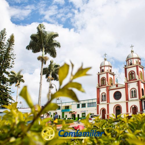 Una colorida iglesia con torres gemelas y adornos rojos se alza detrás de arbustos verdes y palmeras bajo un cielo parcialmente nublado, cerca de la Biblioteca Pública Santa Cecilia. El logotipo de Comfamiliar aparece en la parte inferior de la imagen.