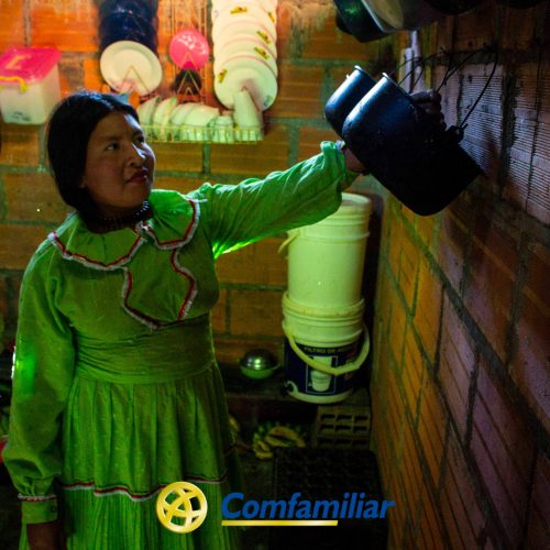 Una mujer con un vestido verde brillante busca una tetera negra en una pared de ladrillo de una cocina poco iluminada del Corregimiento de Santa Cecilia. En los estantes hay platos y utensilios de cocina. El logotipo de Comfamiliar aparece en la parte inferior de la imagen.