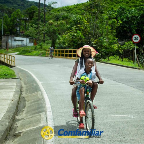 Una mujer y un niño sonríen mientras montan juntos en bicicleta por una carretera de la zona verde y rural de Santa Cecilia. El niño conduce mientras la mujer va sentada detrás de él. Los logotipos de la Biblioteca Pública y Comfamiliar Risaralda son visibles en la parte inferior de la imagen.