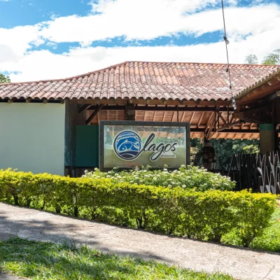 Entrada del restaurante Lagos con un cartel con el logotipo del restaurante, rodeado de vegetación, arbustos y un edificio con tejado de tejas al fondo, bajo un cielo parcialmente nublado.