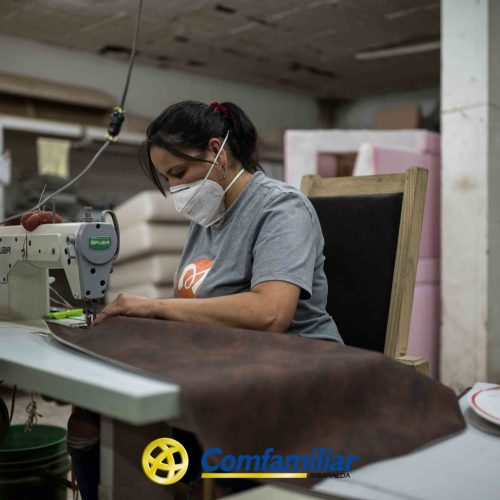 Una mujer con mascarilla utiliza una máquina de coser para coser tela marrón en un taller de la Biblioteca Pública Jaime Fernández Botero. Está concentrada en su trabajo, con rollos de material y espuma cerca. En la parte inferior se ve el logotipo de Comfamiliar.