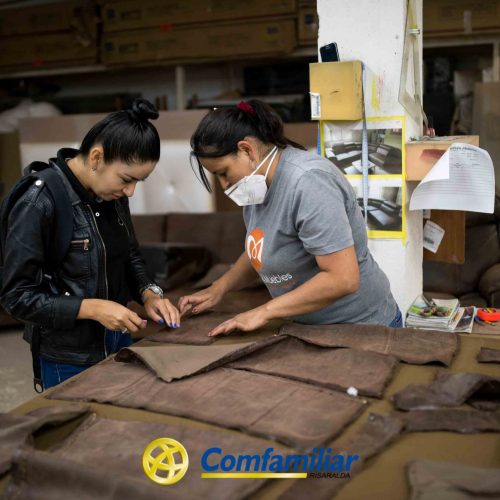 Dos mujeres trabajan juntas, examinando y ordenando trozos de tela marrón sobre una mesa en un taller. Una lleva chaqueta negra, la otra camisa gris y mascarilla. En primer plano se ve el logotipo de Comfamiliar y la Biblioteca Pública Jaime Fernández Botero.