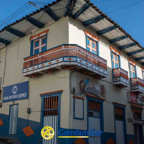 Un edificio tradicional de dos plantas situado en la esquina de una calle, cerca de la Biblioteca Pública León de Greiff, presenta coloridos balcones con detalles en naranja, azul y blanco. En la planta baja se ven varios rótulos comerciales, como "Comfamiliar" y "ELEC".