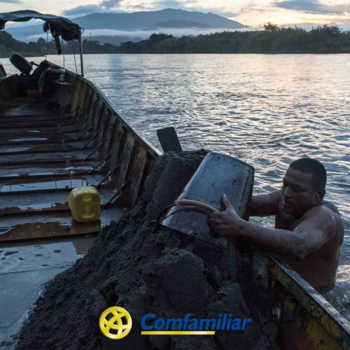 Un hombre se encuentra en un río junto a una barca de madera, levantando un cubo de arena hacia la barca al atardecer. Al fondo se ven montañas y árboles, cerca de la Biblioteca Pública Municipal. El logotipo de Comfamiliar aparece en la parte inferior.