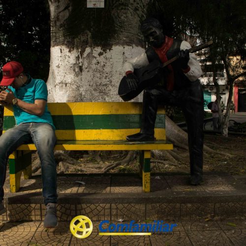 Un hombre con gorra roja se sienta en un banco amarillo y verde junto a la estatua de un guitarrista bajo un gran árbol del parque, cerca de la Biblioteca Pública Municipal Francisco Jaramillo Ochoa, con el logotipo de Comfamiliar visible en el suelo.