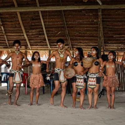 Un grupo de indígenas con atuendos tradicionales y pinturas corporales forman una fila bajo un tejado de paja, algunos con cestas en la mano, y bailan o se preparan para una danza o ceremonia cultural.