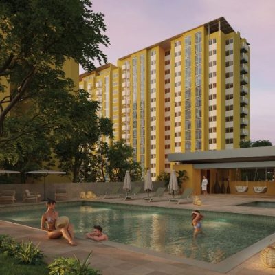 Un moderno complejo de apartamentos, Torres del Bosque, presenta edificios amarillos que rodean una piscina al aire libre. Dos personas se relajan en la piscina mientras otra se sienta en el borde. Tumbonas, sombrillas y vegetación crean un ambiente tranquilo.