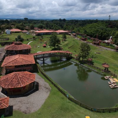 Vista aérea del Parque los arrieros, un parque rural con edificios de tejados rojos, un puente peatonal sobre un estanque, césped verde, árboles y un cielo nublado al fondo. Caminos y vallas dividen el terreno ajardinado.