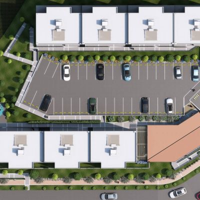 Vista aérea de un moderno complejo residencial en Serranías de Reservado con dos hileras de edificios de tejados blancos, aparcamiento central con coches, zonas verdes, un parque infantil y una puerta de entrada rodeada de caminos ajardinados y árboles.