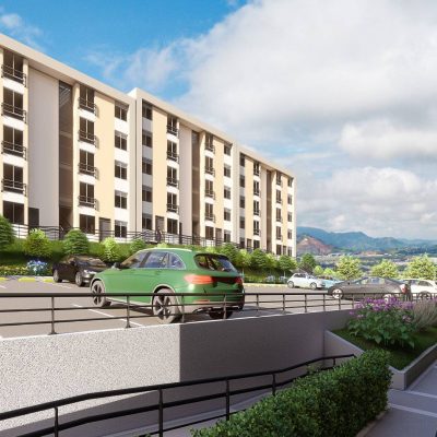Un moderno complejo de apartamentos con edificios de color beige, varias plantas, coches aparcados y verdes jardines, frente a la belleza reservada de las lejanas Serranías bajo un cielo azul parcialmente nublado.