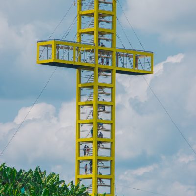 Una alta torre metálica amarilla en forma de cruz se eleva sobre el verde follaje del Parque de las Bailarinas, con gente visible subiendo escaleras y de pie en plataformas de observación. El cielo está parcialmente nublado en el fondo.