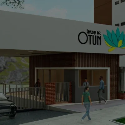 La entrada de un edificio moderno con un cartel en el que se lee Reserva del Otún, gente caminando cerca de la puerta, un coche negro aparcado, árboles y el cercano edificio de apartamentos Riveras del Otún visible al fondo.