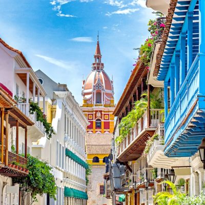 Coloridos edificios de estilo colonial con balcones de madera se alinean en una estrecha calle que conduce a una alta y ornamentada torre de iglesia al fondo, bajo un brillante cielo azul.