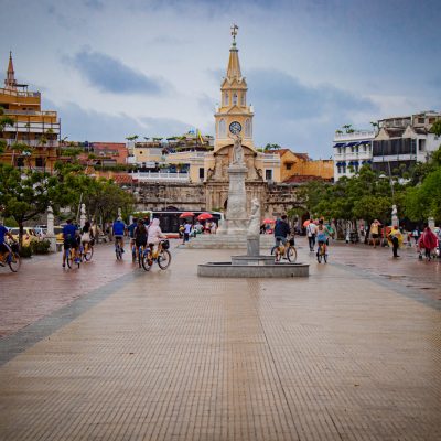 La gente pasea y monta en bicicleta por una amplia plaza con una estatua central y una torre del reloj, rodeada de árboles y edificios de colores bajo un cielo nublado.