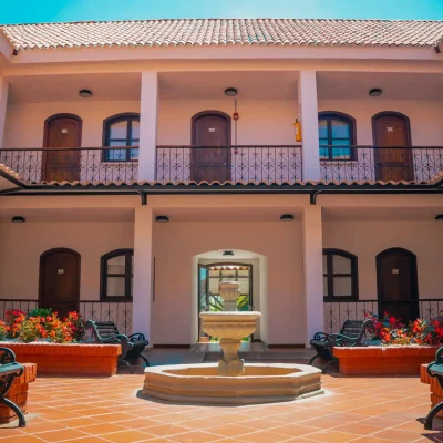 Patio abierto del Hotel Chapola con una fuente central de piedra, rodeada de bancos y parterres. Edificio de dos plantas con puertas de madera arqueadas, barandillas de hierro y tejado de tejas bajo un cielo azul brillante.