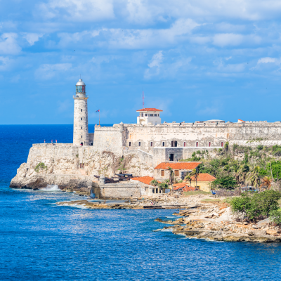 Un faro de piedra y una fortaleza se alzan sobre una costa rocosa de Cuba, con vistas al mar azul, edificios de tejados rojos dispersos, una exuberante vegetación verde y un cielo parcialmente nublado al fondo.