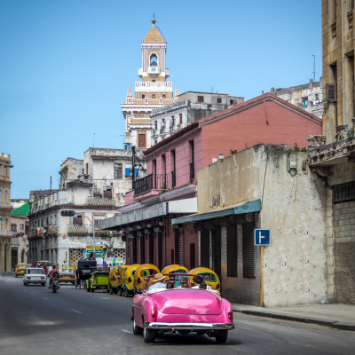 Un coche de época rosa brillante recorre una calle de La Habana, Cuba, pasando junto a edificios antiguos, coloridos tuk-tuks y una alta torre ornamentada bajo un cielo azul despejado.
