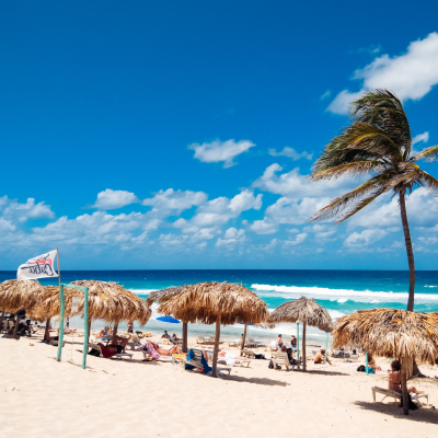 Playa de arena en Cuba con gente descansando bajo sombrillas de paja, una palmera inclinada, una bandera de salvavidas y olas turquesas bajo un cielo azul brillante con nubes blancas dispersas: perfecto para tu próximo plan bajo.