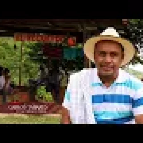 Un hombre con polo a rayas y gorra blanca se sienta al aire libre con un paño al hombro, cerca de la rústica estructura de madera de la Biblioteca Pública Municipal Juan XXIII, rodeada de vegetación.