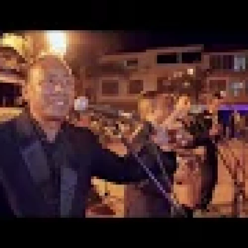 Un sonriente músico con traje negro canta ante un micrófono durante una actuación nocturna al aire libre en la Biblioteca Pública Comfamiliar Guática, con una banda y un público visibles al fondo.