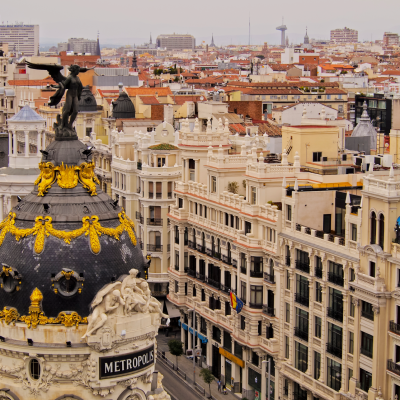 Vista aérea del edificio Metrópolis, con su cúpula negra ornamentada y su estatua alada, rodeado de edificios históricos y calles del centro de Madrid, España.