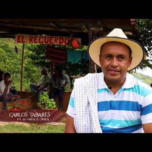 Un hombre con sombrero blanco y vestimenta tradicional se sienta al aire libre frente a una estructura rústica de madera con un cartel en el que se lee El Recuerdo, cerca de la Biblioteca Municipal. Varias personas se reúnen bajo la sombra. El texto dice: Carlos Tabares.