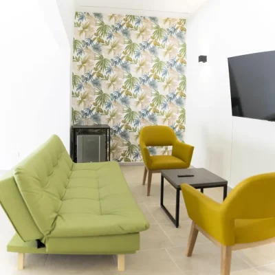 El moderno salón de Cabañas Consotá cuenta con un futón verde, dos sillas amarillo mostaza, una mesa de centro rectangular de madera, un televisor montado en la pared y una pared de acento con motivos de hojas tropicales.