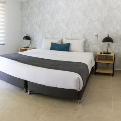 Un dormitorio moderno en Cabañas Consotá cuenta con una cama grande con ropa de cama blanca, una manta gris oscuro, dos mesillas de noche con lámparas negras y una ventana equipada con persianas de doble capa que dejan entrar abundante luz natural.